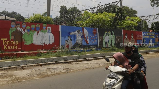 Pengendara motor melintas di depan mural tentang pandemi COVID-19 di Bukit Duri, Jakarta, Selasa (20/10/2020). [Suara.com/Angga Budhiyanto]