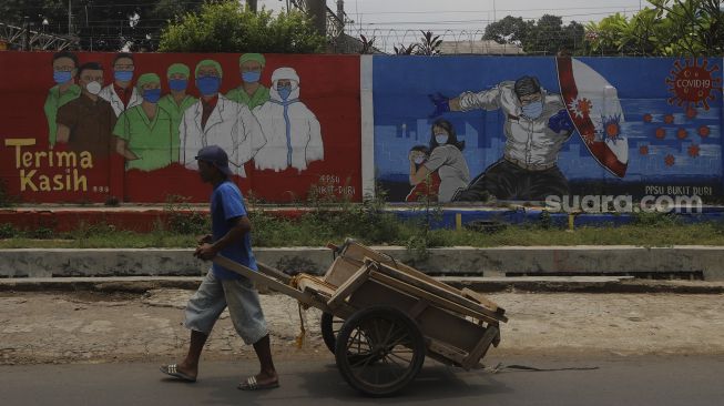Pekerja melintas di depan mural tentang pandemi COVID-19 di Bukit Duri, Jakarta, Selasa (20/10/2020). [Suara.com/Angga Budhiyanto]