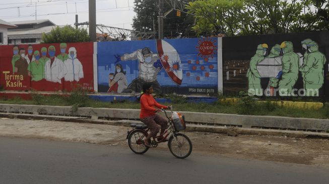 Pesepeda melintas di depan mural tentang pandemi COVID-19 di Bukit Duri, Jakarta, Selasa (20/10/2020). [Suara.com/Angga Budhiyanto]