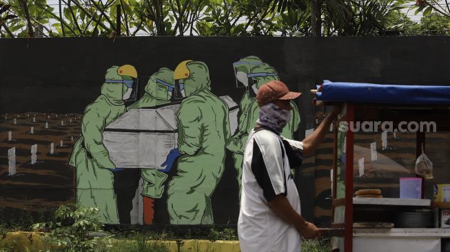 Pedagang melintas di depan mural tentang pandemi COVID-19 di Bukit Duri, Jakarta, Selasa (20/10/2020). [Suara.com/Angga Budhiyanto]