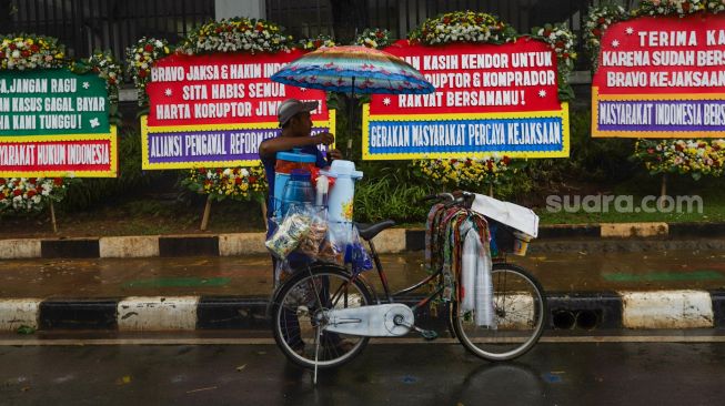 Pedagang kopi keliling melayani pembeli didepan karangan bunga terkait kasus korupsi Jiwasraya didepan Kejaksaan Agung RI, Jakarta Selatan, Selasa (27/10). [Suara.com/Alfian Winanto]