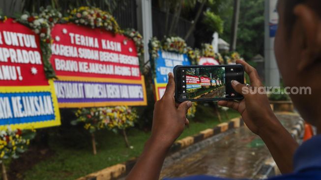 Seorang warga mengabadikan dengan smartphonenya karangan bunga terkait kasus korupsi Jiwasraya didepan Kejaksaan Agung RI, Jakarta Selatan, Selasa (27/10). [Suara.com/Alfian Winanto]