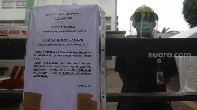 Petugas berjaga di dekat kertas pemberitahuan penutupan sementara Puskesmas Duren Sawit, Jakarta Timur, Rabu (6/1/2021). [Suara.com/Angga Budhiyanto]