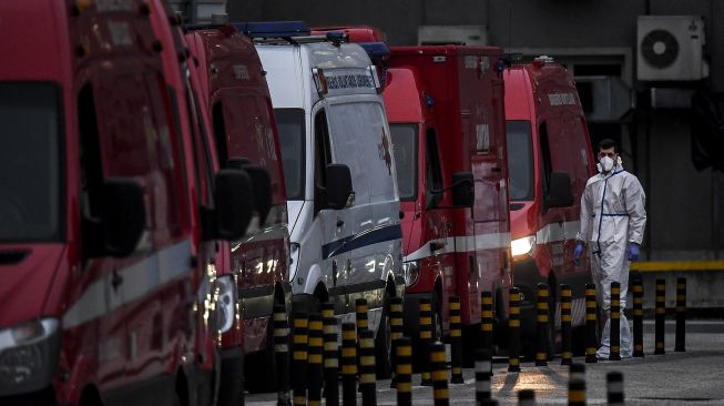 Seorang petugas kesehatan yang mengenakan pakaian pelindung berdiri di samping lusinan ambulans yang menunggu di luar layanan darurat COVID-19 di Rumah Sakit Santa Maria, Lisbon, pada (28/1/2021). [PATRICIA DE MELO MOREIRA / AFP]