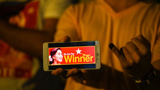 Seorang pendukung partai Liga Nasional untuk Demokrasi (NLD) memegang ponsel yang menunjukkan gambar Aung San Suu Kyi di depan markas partai di Yangon pada 8 November 2020, [Ye Aung THU / AFP]