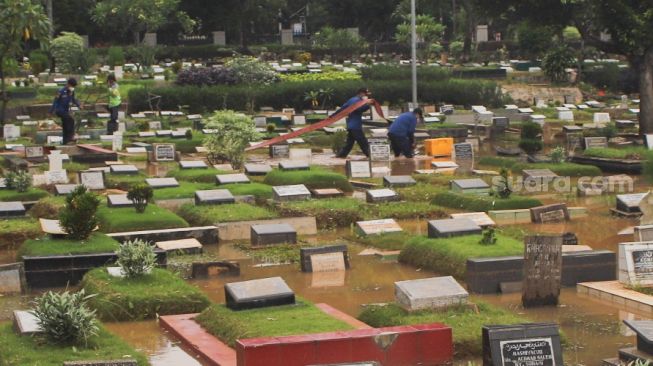 Petugas membawa alat untuk menyedot air di area pemakaman yang terendam banjir di TPU Karet Bivak, Jakarta Pusat, Minggu (7/2/2021). [Suara.com/Alfian Winanto]