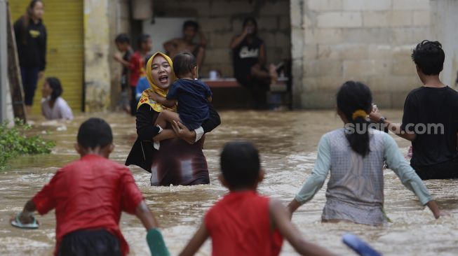 Seorang warga menggendong anaknya berjalan menerjang banjir yang merendam Kelurahan Cipinang Melayu, Jakarta, Jumat (19/2/2021). [Suara.com/Angga Budhiyanto]