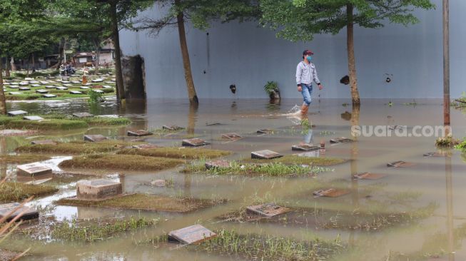 Warga melintas di sekitar area makam yang terendam banjir di TPU Jeruk Purut, Jakarta Selatan, Sabtu (20/2/2021). [Suara.com/Alfian Winanto]