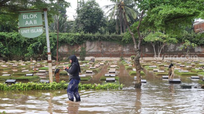 Warga melintas di sekitar area makam yang terendam banjir di TPU Jeruk Purut, Jakarta Selatan, Sabtu (20/2/2021). [Suara.com/Alfian Winanto]