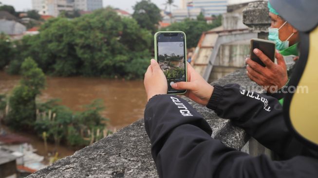 Seorang pengendara motor mengambil gambae dengan smartphone saat menonton banjir dan aliran kali Ciliwung yang meluap dari atas Flyover Kalibata, Jakarta Selatan, Sabtu (20/2/2021). [Suara.com/Alfian Winanto]