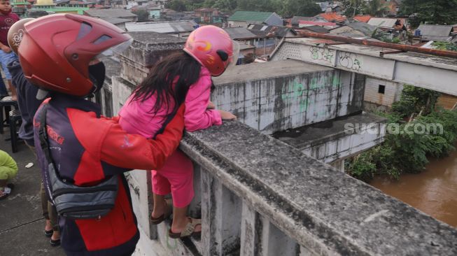 Seorang pengendara motor mengajak anaknya untuk menonton banjir dan aliran kali Ciliwung yang meluap dari atas Flyover Kalibata, Jakarta Selatan, Sabtu (20/2/2021). [Suara.com/Alfian Winanto]