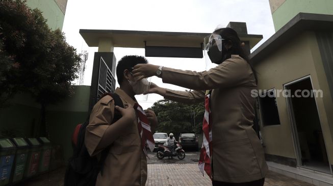 Seorang Guru membantu memakaikan pelindung wajah kepada murid pada hari pertama uji coba pembelajaran tatap muka di SD Negeri Kenari 08 Pagi, Jakarta, Rabu (7/4/2021). [Suara.com/Angga Budhiyanto]
