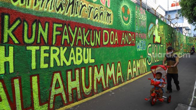 Warga berjalan di depan mural bernuansa Islami di Gang Pelangi, Kalibata, Jakarta Selatan, Sabtu (17/4/2021). [Suara.com/Alfian Winanto]