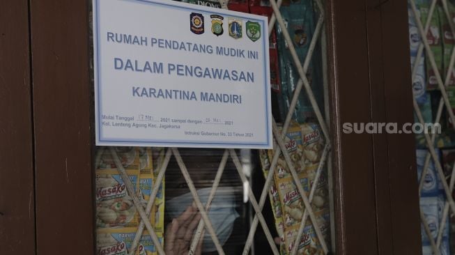 Seorang warga yang baru kembali ke Jakarta berdiri di balik stiker tanda menjalani isolasi mandiri di rumahnya di kawasan Lenteng Agung, Jakarta, Senin (17/5/2021). [Suara.com/Angga Budhiyanto]