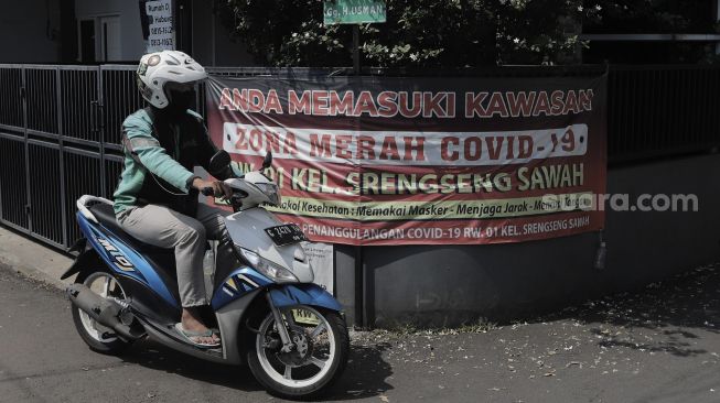 Pengendara sepeda motor melintasi spanduk penanda zona merah di Jalan H Shibi II, Kelurahan Srengseng Sawah, Jagakarsa, Jakarta Selatan, Jumat (28/5/2021). [Suara.com/Angga Budhiyanto]