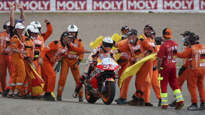 Pembalap Honda Spanyol Marc Marquez (tengah) disambut oleh staf sirkuit dengan plakat bertuliskan nomor '11' setelah memenangkan Grand Prix MotoGP Jerman di sirkuit balap Sachsenring, Hohenstein-Ernstthal, Jerman Timur, pada (20/6/2021). [Ronny HARTMANN / AFP]