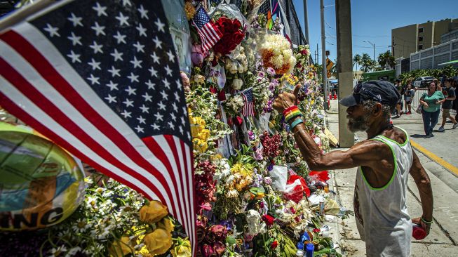 Orang-orang menambahkan bunga ke tugu peringatan bergambar beberapa orang yang hilang dari gedung kondominium Champlain Towers Sout yang sebagian runtuh di Surfside, Florida, pada (3/7/2021). [Giorgio Viera / AFP]