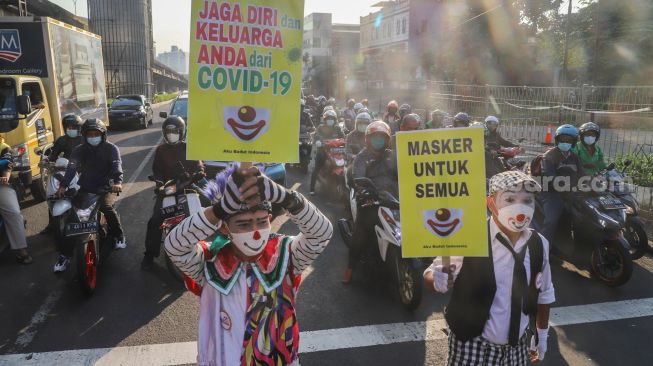 Angggota Komunitas Aku Badut Indonesia (ABI) membawa poster berisi ajakan untuk menggunakan masker di kawasan Cilandak, Jakarta Selatan, Senin (12/7/2021). [Suara.com/Alfian Winanto]