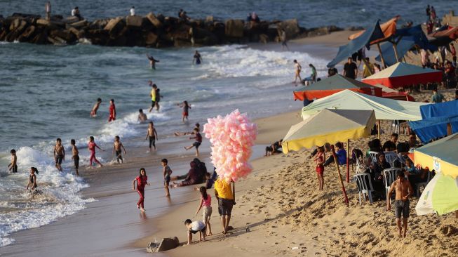 Seorang pedagang berjalan di pantai ketika orang-orang bersantai saat musim panas yang tinggi di Gaza, Palestina, pada (16/7/2021). [MOHAMMED ABED / AFP]