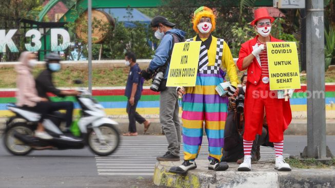 Angggota Komunitas Aku Badut Indonesia (ABI) melakukan aksi kampanye untuk mengajak masyarakat melakukan vaksinasi dan mematuhi protokol kesehatan di Kawasan Juanda, Depok, Jawa Barat, Jumat (23/7/2021). [Suara.com/Alfian Winanto]