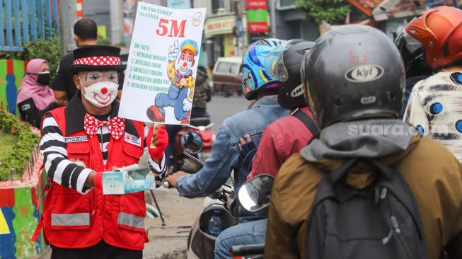 Angggota Komunitas Aku Badut Indonesia (ABI) membagikan masker kepada pengendara motor saat melakukan aksi kampanye untuk mengajak masyarakat melakukan vaksinasi dan mematuhi protokol kesehatan di Kawasan Juanda, Depok, Jawa Barat, Jumat (23/7/2021). [Suara.com/Alfian Winanto]