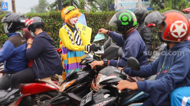 Angggota Komunitas Aku Badut Indonesia (ABI) membagikan masker kepada pengendara motor saat melakukan aksi kampanye untuk mengajak masyarakat melakukan vaksinasi dan mematuhi protokol kesehatan di Kawasan Juanda, Depok, Jawa Barat, Jumat (23/7/2021). [Suara.com/Alfian Winanto]