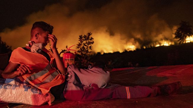 Seorang sukarelawan beristirahat dengan latar belakang api yang membubung dari hutan yang terbakar di desa Akcayaka, Milas, Provinsi Mugla, Turki, pada (6/8/2021). [Yasin AKGUL / AFP]