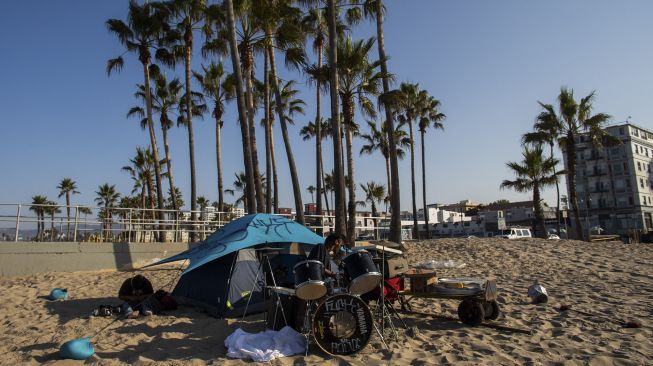 Seorang tunawisma bermain drum pada pagi hari di Pantai Venesia, California, Amerika Serikat, pada (12/8/2021). [Apu GOMES / AFP]