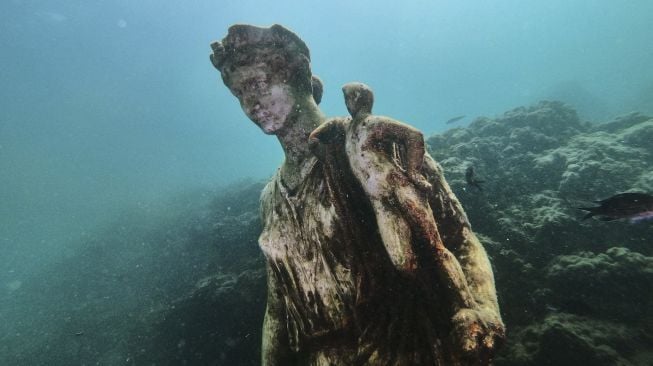 Ikan berenang melewati salinan patung asli yang disimpan di Museum Baiae, kota Romawi kuno Baiae yang tenggelam  di Taman Bawah Air Baiae, bagian dari situs kompleks Taman Arkeologi Campi Flegrei di Pozzuoli dekat Napoli, Italia, pada (18/8/2021). [Andreas SOLARO / AFP]