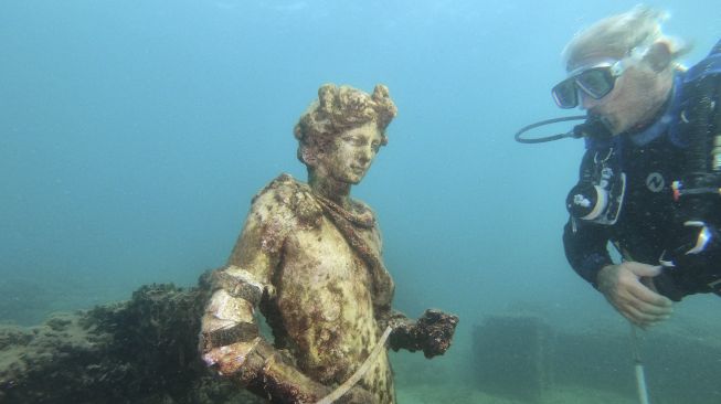 Pemandu selam menunjukkan kepada wisatawan salinan patung asli yang disimpan di Museum Baiae, kota Romawi kuno Baiae yang tenggelam di Taman Bawah Air Baiae, bagian dari situs kompleks Taman Arkeologi Campi Flegrei di Pozzuoli dekat Napoli, Italia, pada (18/8/2021). [Andreas SOLARO / AFP]