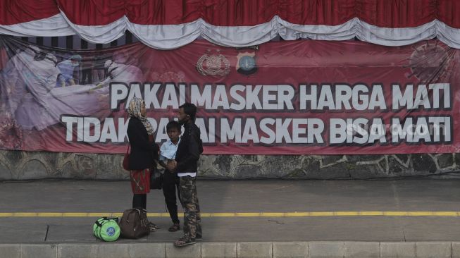 Warga berdiri di depan spanduk berisi imbauan untuk memakai masker yang terpasang di Jalan Gatot Subroto, Jakarta, Senin (23/8/2021). [Suara.com/Angga Budhiyanto]