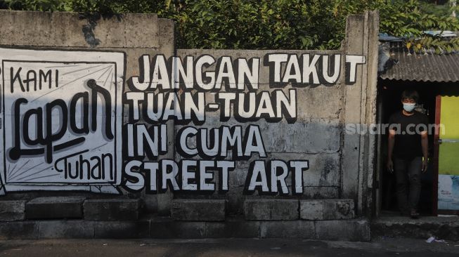 Warga berdiri di dekat mural yang bertuliskan "jangan takut tuan-tuan ini cuma street art" di Jakarta, Rabu (25/8/2021). [Suara.com/Angga Budhiyanto]