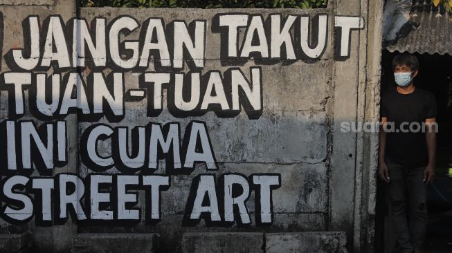 Warga berdiri di dekat mural yang bertuliskan "jangan takut tuan-tuan ini cuma street art" di Jakarta, Rabu (25/8/2021). [Suara.com/Angga Budhiyanto]