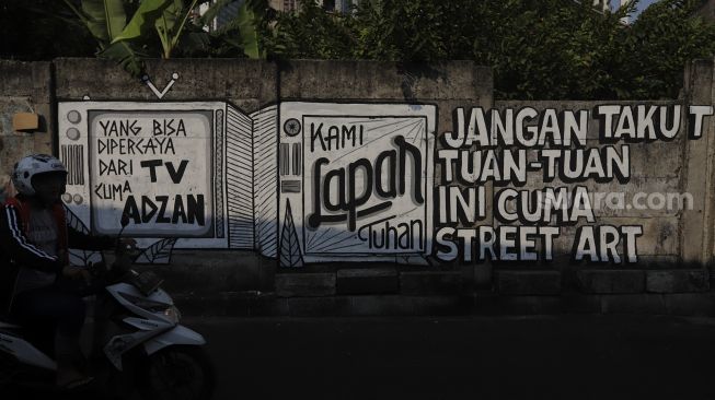 Pengendara sepeda motor melintas di dekat mural yang bertuliskan "jangan takut tuan-tuan ini cuma street art" di Jakarta, Rabu (25/8/2021). [Suara.com/Angga Budhiyanto]