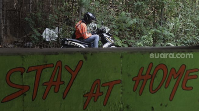 Seorang pengendara sepeda motor berhenti di dekat mural bertema COVID-19 di kolong Jalan Tol Lingkar Luar Jakarta, Cipayung, Jakarta Timur, Jumat (3/9/2021). [Suara.com/Angga Budhiyanto]