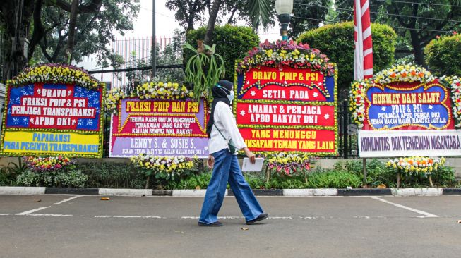 Warga melintasi karangan bunga yang terpajang di halaman kantor DPRD DKI Jakarta, Jakarta Pusat, Jumat (3/9/2021). [Suara.com/Alfian Winanto]