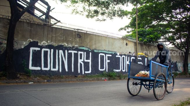 Pengendara becak tengah melintas di mural bertuliskan “ Country of Jokes” di Tangerang, Jumat (3/9).  ( Suara.com/Hilal Rauda Fiqry) 