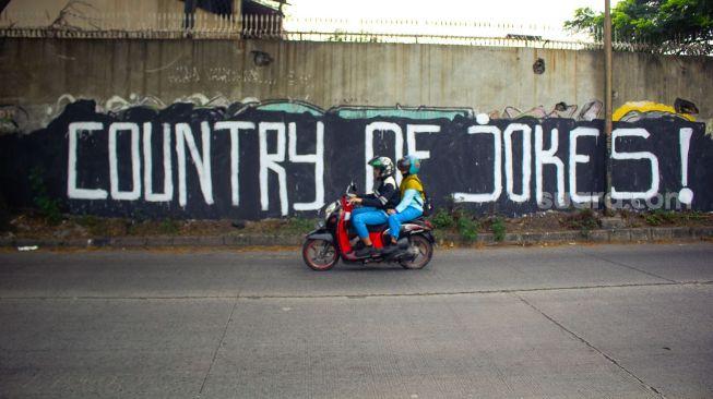 Pengendara sepeda motor tengah melintas di mural bertuliskan “ Country of Jokes” di Tangerang, Jumat (3/9).  ( Suara.com/Hilal Rauda Fiqry) 