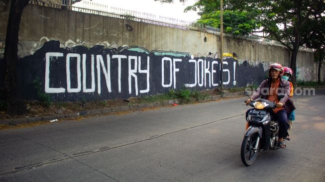 Pengendara sepeda motor tengah melintas di mural bertuliskan “ Country of Jokes” di Tangerang, Jumat (3/9).  ( Suara.com/Hilal Rauda Fiqry) 