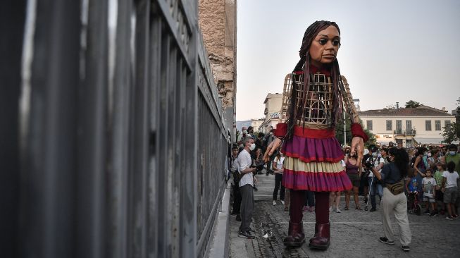 "Little Amal", boneka raksasa yang menggambarkan seorang gadis pengungsi muda berdiri di dekat pagar saat diarak di beberapa jalan di Athena, Yunani, pada (2/9/2021). [Louisa GOULIAMAKI / AFP]