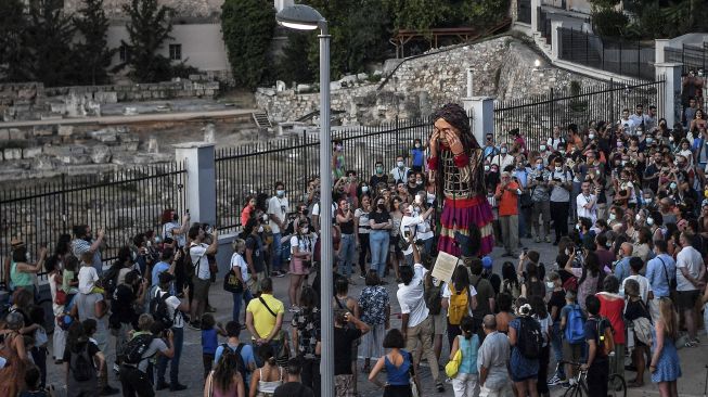 Warga berkumpul di sekitar "Little Amal", boneka raksasa yang menggambarkan seorang gadis pengungsi muda saat diarak di beberapa jalan di Athena, Yunani, pada (2/9/2021). [Louisa GOULIAMAKI / AFP]