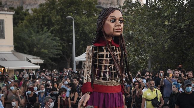 Warga berkumpul di sekitar "Little Amal", boneka raksasa yang menggambarkan seorang gadis pengungsi muda saat diarak di beberapa jalan di Athena, Yunani, pada (2/9/2021). [Louisa GOULIAMAKI / AFP]