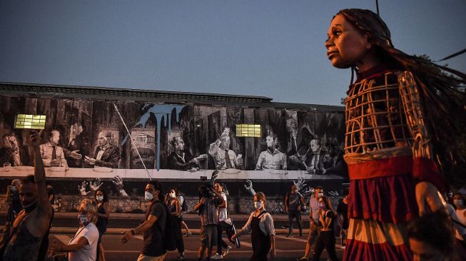 Warga berkumpul di sekitar "Little Amal", boneka raksasa yang menggambarkan seorang gadis pengungsi muda saat melewati mural karya seniman jalanan Yunani INO, saat diarak di beberapa jalan di Athena, Yunani, pada (2/9/2021). [Louisa GOULIAMAKI / AFP]