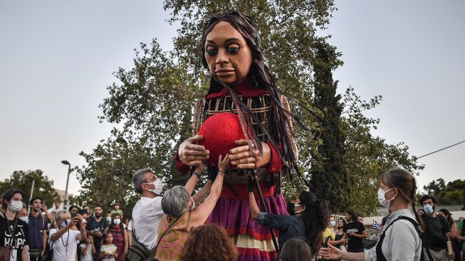 Warga memberikan Benang Ariadne untuk menemukan jalan kepada "Little Amal", boneka raksasa yang menggambarkan seorang gadis pengungsi muda saat diarak di beberapa jalan di Athena, Yunani, pada (2/9/2021). [Louisa GOULIAMAKI / AFP]