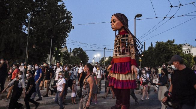Warga berkumpul di sekitar "Little Amal", boneka raksasa yang menggambarkan seorang gadis pengungsi muda saat diarak di beberapa jalan di Athena, Yunani, pada (2/9/2021). [Louisa GOULIAMAKI / AFP]