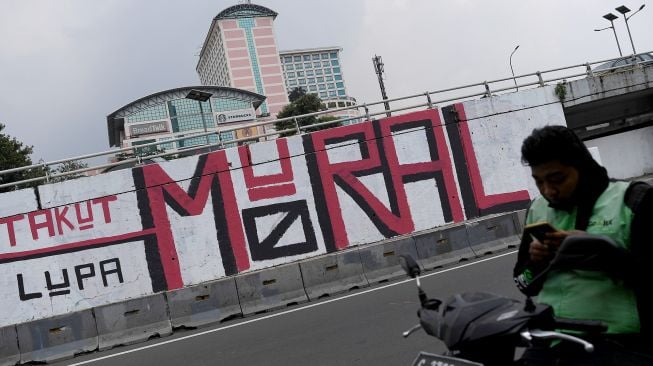 Pengendara ojek daring menunggu penumpang di depan mural yang berisi kritikan di Jakarta Barat, Minggu (19/9/2021).  ANTARA FOTO/Sigid Kurniawan