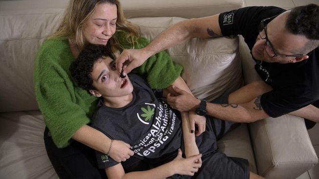 Gabriel Guerra (tengah) digendong oleh ibunya, Vanessa Opitz, sementara ayahnya Ricardo Guerra memberinya ganja obat di Rio de Janeiro, Brasil, pada (20/9/2021). [Carl de Souza / AFP]