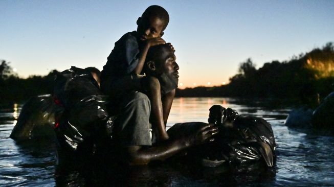 Seorang pria menggendong seorang anak di pundaknya saat para migran Haiti menyeberangi Sungai Rio Grande di perbatasan Meksiko-AS, pada (23/9/2021). [PEDRO PARDO / AFP]