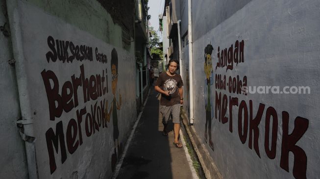 Warga berjalan melintasi mural larangan merokok di RW.06 Kelurahan Kayu Manis, Matraman, Jakarta, Jumat (8/10/2021). [Suara.com/Angga Budhiyanto]