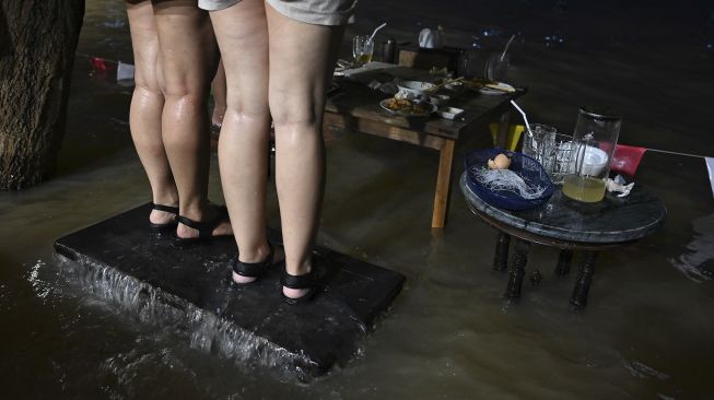 Pengunjung berdiri di kursi mereka sambil menikmati makan malam di Kafe Antik Chaopraya, saat air banjir dari Sungai Chao Phraya mengalir ke restoran di provinsi Nonthaburi, Bangkok, Thailand, pada (7/10/2021). [Lillian SUWANRUMPHA / AFP]